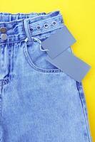 Flat lay, detail of denim jeans with blank gray label on yellow background. photo