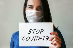 un joven mujer en un médico máscara con un papel en su mano con el inscripción detener covid-19 . foto