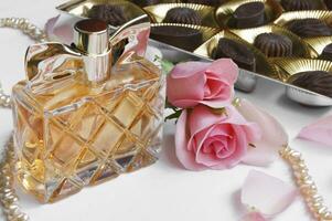 The perfume bottle with rose, rose petals, pearls and chocolates on a light background. photo