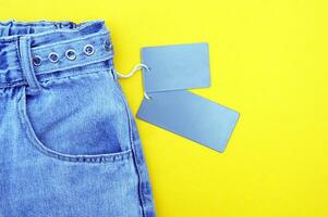 Flat lay, detail of denim jeans with blank gray label on yellow background. photo