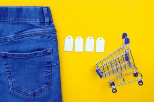 Flat lay, detail of blue jeans, shopping cart with blank white labels on yellow background. photo