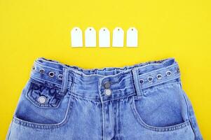 Flat lay, detail of denim jeans with blank white labels on yellow background. photo