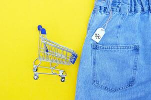 Flat lay, detail of blue jeans, shopping cart with white label on yellow background. photo