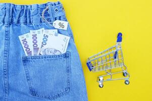 Detail of denim jeans with Ukrainian hryvnia bills in pocket, shopping cart with white label on yellow background. photo