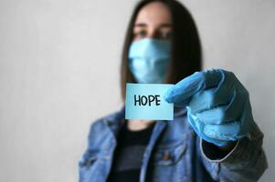 un joven mujer en un médico máscara y guantes, ella sostiene un pegatina con el palabras esperanza. foto