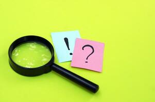 Top view photo of a magnifying glass and sticker with question mark and an exclamation mark lie on a green background.