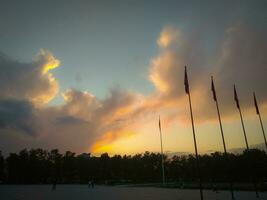 Colorful sunlight of sunset sky heavenly shining above the tree tops and the town square with flags on poles, bottom up view photo