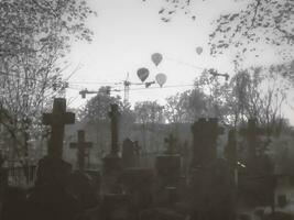 Dramatic black and white artistic photograph of crosses of a graveyard and air balloons flying over it and crawler cranes. photo