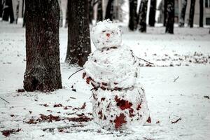 Old dirty snowman with carrot nose covered in fallen brown leaves standing between the trees photo