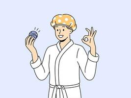 Man with bath bomb is dressed in bathrobe and bandage to protect hair from moisture. Young guy shows OK gesture recommending use of aromatic bath bomb while performing hygiene procedures. vector