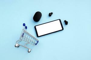 Shopping cart with a smartphone and black wireless headphones on a blue background. photo