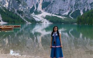 contento asiático mujer turista en pie en el costa de braies lago. dolomitas, Italia. paisaje de famoso lago con hermosa reflexión en agua, árboles, cielo con nubes de madera barco cabaña. viajar. foto