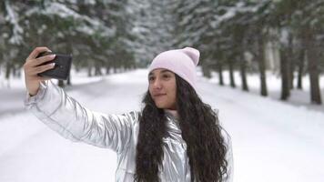 joven hermosa niña hace selfie utilizando un teléfono inteligente mientras en pie en el invierno bosque. lento movimiento hd video