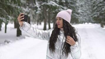 ung skön flicka gör selfie använder sig av en smartphone medan stående i de vinter- skog. långsam rörelse video