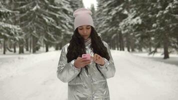 un attraente malto ragazza con un' bene umore va attraverso il inverno foresta nel il pomeriggio e usi un' smartphone. 4k video