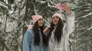 Two beautiful girlfriends make selfie on the background of the winter forest using a smartphone. 4K video