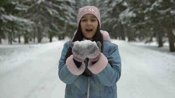 atractivo niña soplo nieve desde manos y sonriente mientras en pie en el invierno bosque en el antecedentes de arboles video