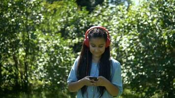 un attrayant Jeune fille avec longue cheveux balades par le parc et écoute à la musique sur casque.lent mouvement. HD video