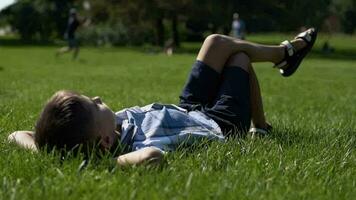 A small attractive boy lies in a park on the grass having a good mood during the day in sunny weather. slow motion. HD. video