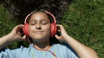 Attractive young girl with long black hair lies on the grass in the park and listens to music in red headphones having a good mood.slow motion. HD video