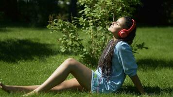 atractivo joven niña con largo negro pelo escuchando a música en auriculares utilizando teléfono inteligente sentado en césped en parque en soleado clima. 4k video
