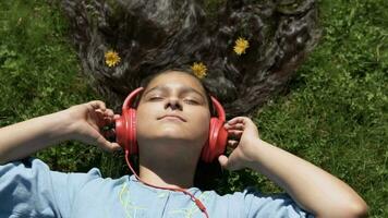 atractivo joven niña con largo negro pelo mentiras en el césped en el parque y escucha a música en auriculares.lento movimiento. video