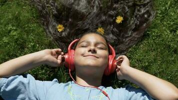 atractivo joven niña con largo pelo mentiras en césped en parque y escucha a música en auriculares.lento movimiento. hd video