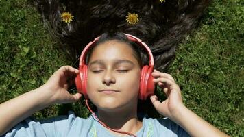 atraente jovem menina com grandes Preto cabelo mentiras em a Relva dentro a parque e escuta para música em fones de ouvido tendo uma Boa humor.lento movimento. video