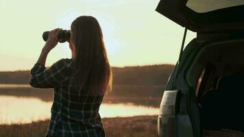 Finding strategy, Explore area, Find solution, Woman tourist. Woman looking through binoculars while standing by the water next to the car during dawn video