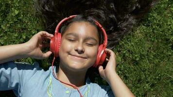 atraente jovem menina com grandes Preto cabelo mentiras em a Relva dentro a parque e escuta para música dentro vermelho fones de ouvido tendo uma Boa humor.lento movimento. video