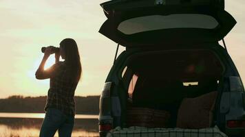 achando estratégia, explorar área, encontrar solução, mulher turista. mulher olhando através binóculos enquanto em pé de a água Próximo para a carro durante pôr do sol video