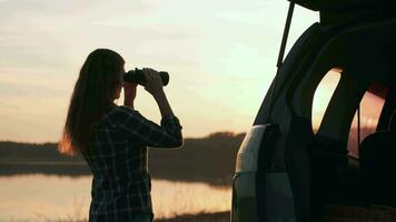 achando estratégia, explorar área, encontrar solução, mulher turista. mulher turista olhando através binóculos enquanto em pé de a água Próximo para a carro durante pôr do sol video