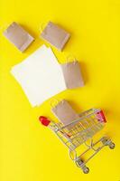 Mini trolley with small craft bags and blank white paper for lettering or shopping list on yellow background. photo