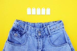 Flat lay, detail of denim jeans with blank white labels on yellow background. photo