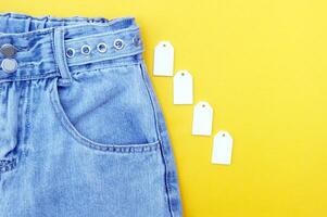 Flat lay, detail of denim jeans, blank white labels on yellow background. photo