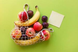 Red apples, bananas, plums, string bag and blank paper on a bright green background photo