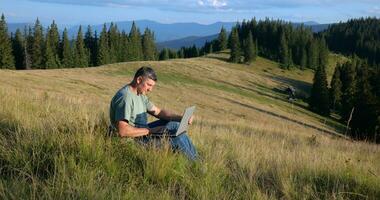 uma homem senta em uma lindo Prado dentro a montanhas, trabalho em uma computador portátil. conceito do freelancer, digital nômade ou controlo remoto escritório. video