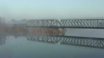 antenne visie van een trein op reis over- een spoorweg brug over- een rivier- in de vroeg ochtend. een locomotief met rijtuigen beweegt langs een smalspoor spoorweg video
