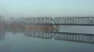 Aerial view of a train traveling over a railway bridge over a river in the early morning. A locomotive with carriages moves along a narrow-gauge railway video