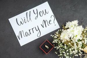 Top View Engagement Ring in Red Box Will You Marry Me Written on Paper Flowers on Table photo