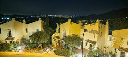 Nighttime view of a residential area with illuminated houses and surrounding trees against a dark mountainous backdrop. photo