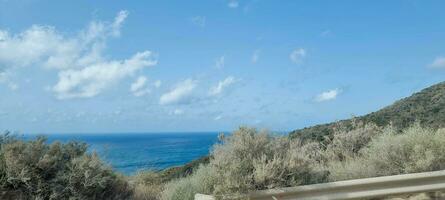 Coastal landscape with clear blue sky, fluffy clouds, and a view of the sea from a roadside. photo