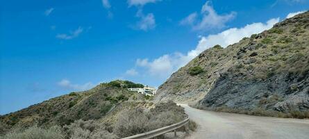 Winding mountain road with clear blue sky and sparse vegetation leading to a distant white building. photo