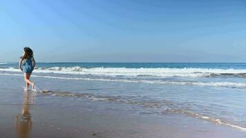 Full length portrait of a happy little child girl playing barefoot on the tropical beach, enjoying the sea water splashing on her feet, running leaving footsteps washed by waves on the sandy beach video