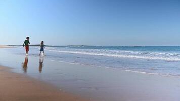 Caucasian beautiful kids walking together barefoot and playing on the empty sandy beach, smiling and having fun, standing by the water, enjoying how the waves wash their legs while splashing on sand video