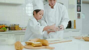 joven asiático padre y su hijo vistiendo cocinero uniforme horneando juntos en cocina a hogar video