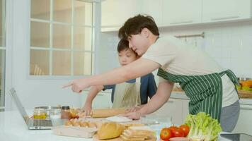 Happy young Asian father and kid are using a laptop computer to learn how to cooking food in their home kitchen. video