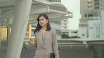 un joven hermosa mujer de negocios es Bebiendo café caminando en moderno ciudad , negocio tecnología , ciudad estilo de vida concepto video