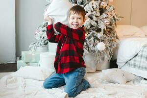 A boy in a red checkered shirt sits by the Christmas tree with gifts on the floor and fights with a pillow photo