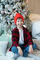 A boy in a red hat and a plaid shirt sits near a Christmas tree with gifts on the floor and smiles photo